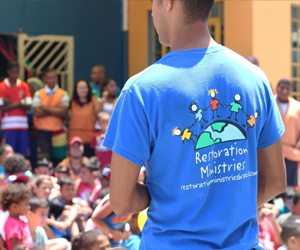 Man wearing a Restoration Ministries shirt standing in front of a group of children