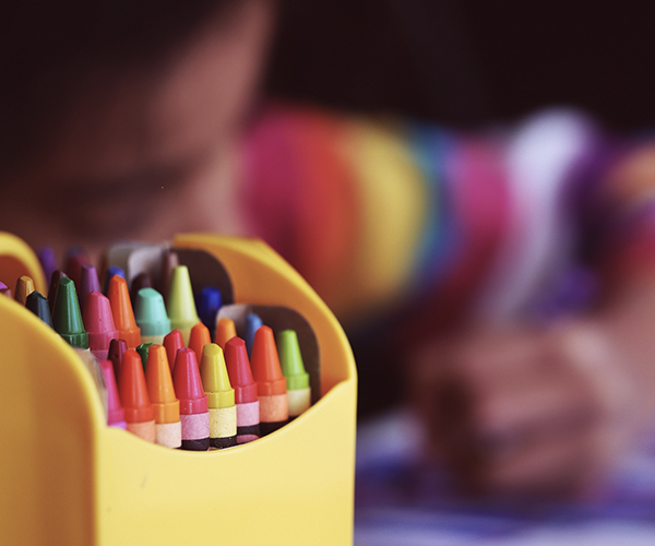 A box of crayons with a young child coloring in the background